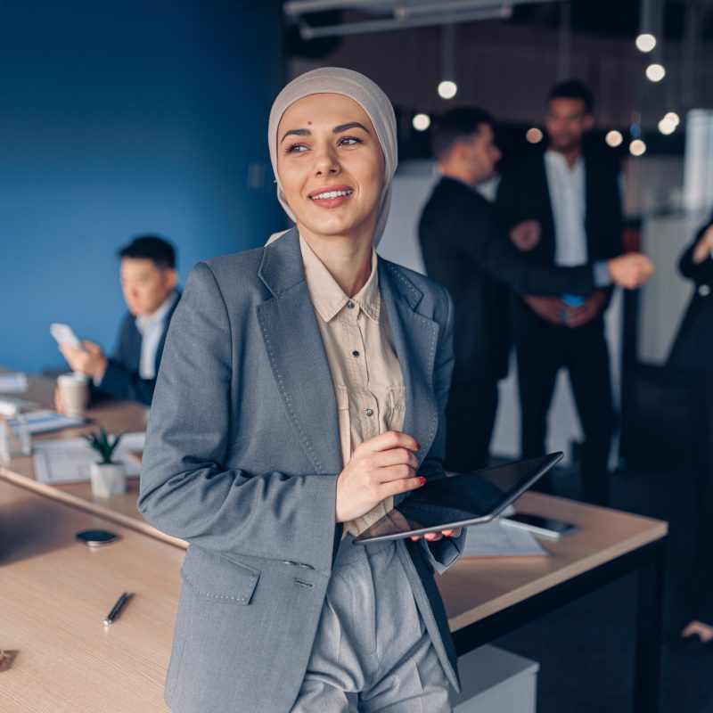 Muslim businesswoman in hijab looking at side working on digital tablet on colleagues background